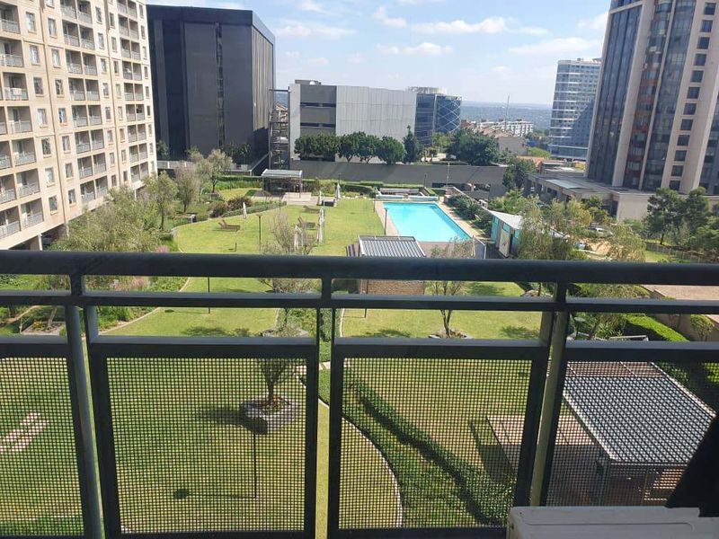 Elevated view from a balcony overlooking a well-maintained residential complex garden, swimming pool area, and surrounding urban high-rise buildings.