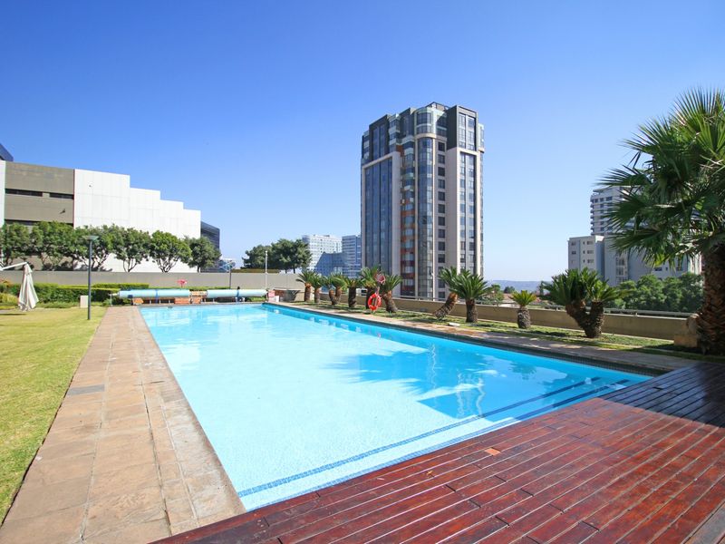 A well-maintained outdoor swimming pool area featuring timber decking, stone paving, and landscaped garden surrounds within an apartment complex.