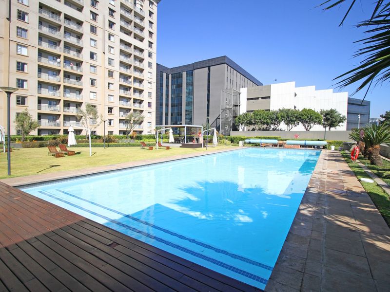 A well-maintained communal swimming pool area featuring timber decking, stone paving, and landscaped lawn within an apartment complex.