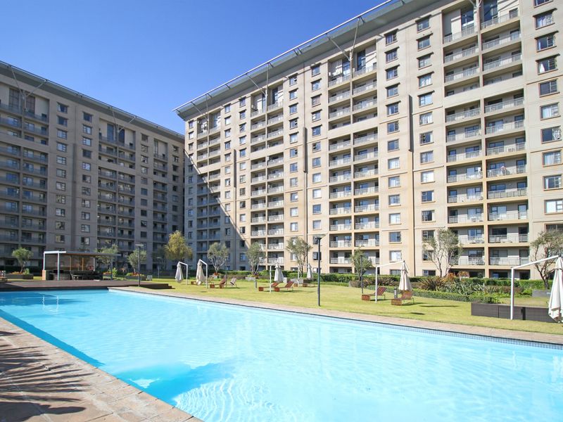 A well-maintained communal swimming pool area set within a landscaped garden, surrounded by large apartment buildings.