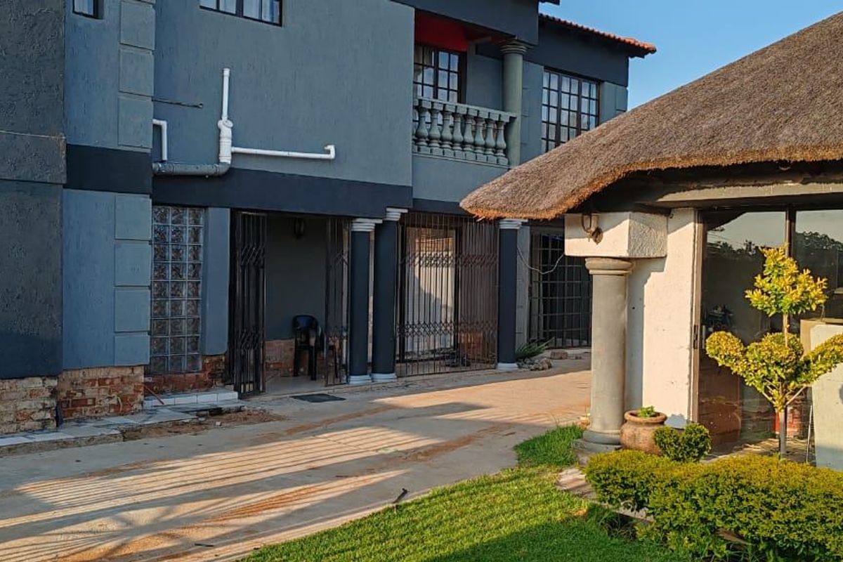 Exterior view of a multi-story house featuring a thatched-roof lapa, paved driveway, and covered patio area with security gates.