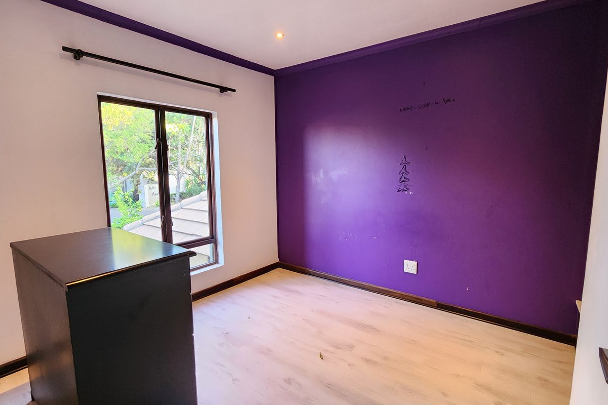 An empty bedroom featuring light-toned laminate flooring, a window with a curtain rod, and a prominent purple accent wall requiring cosmetic attention.