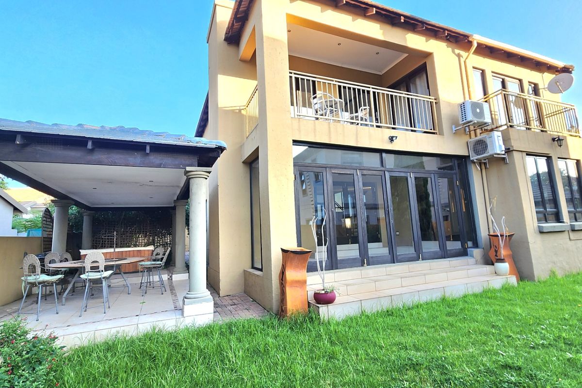 Rear view of a two-story house featuring a covered patio, upper balcony, bi-fold doors, and external air conditioning units set against a lawn.