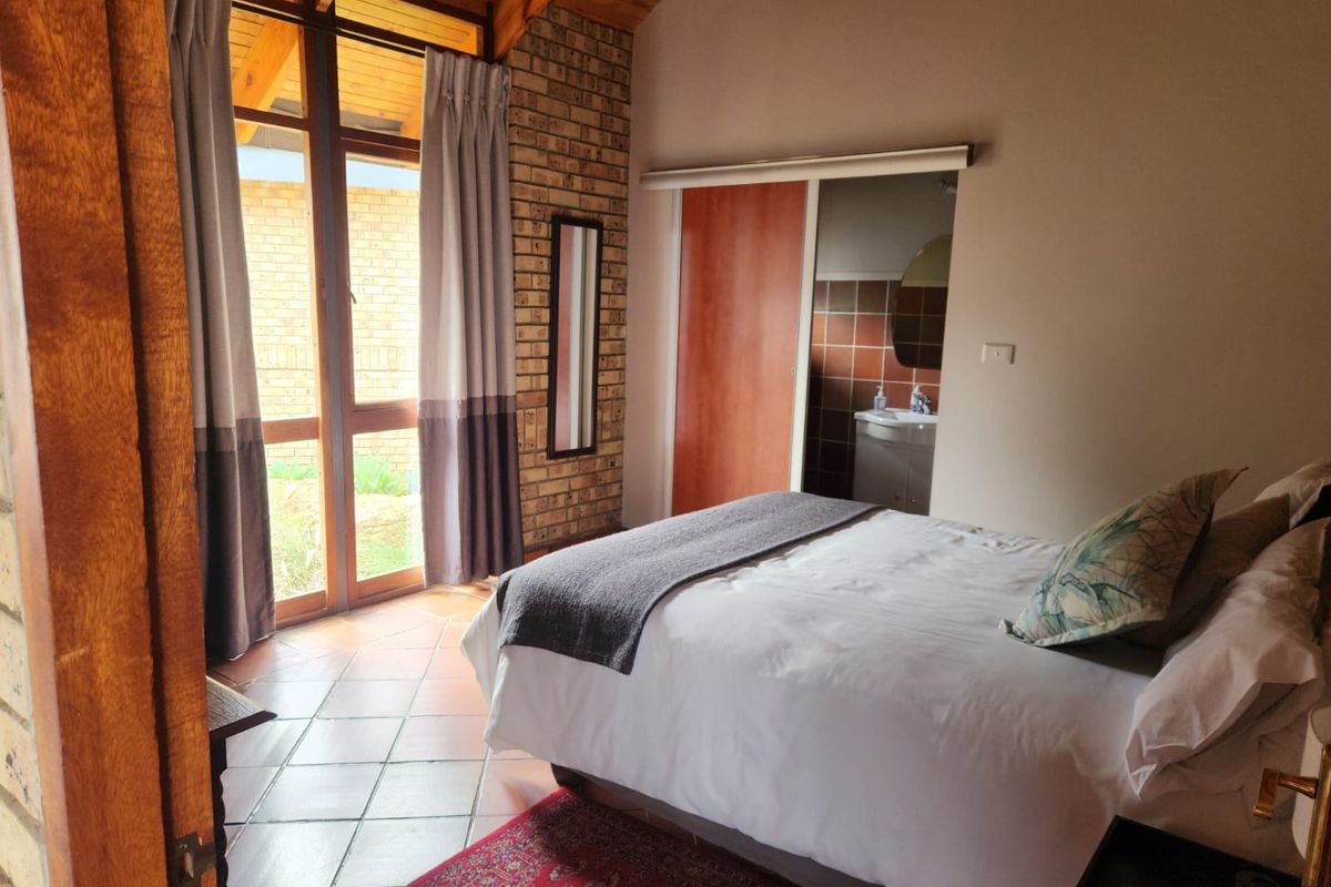 A bedroom featuring a high exposed timber ceiling, facebrick wall, and terracotta tiles, with direct access to an en-suite bathroom and outdoor area.