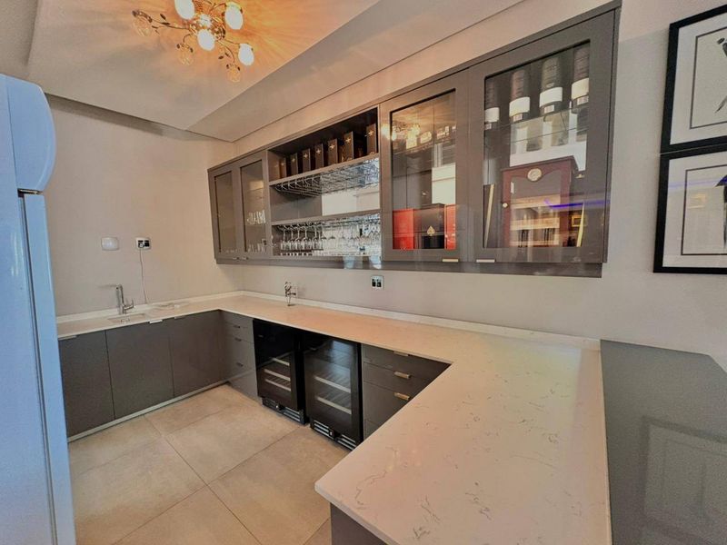 A modern, well-appointed home bar featuring sleek grey cabinetry, stone countertops, integrated wine coolers, and professional-style glass display shelving.