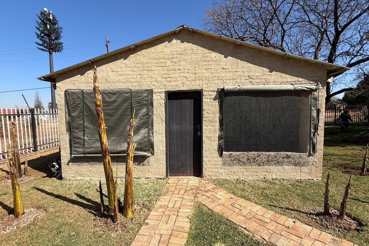 A detached outbuilding featuring a brick facade, central entrance door, and protective window screens, set within a garden area with a brick pathway.
