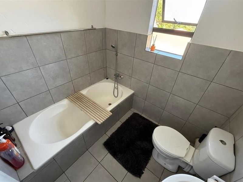 A functional bathroom featuring a built-in bathtub with a handheld shower mixer, a standard toilet, and neutral grey wall and floor tiling.