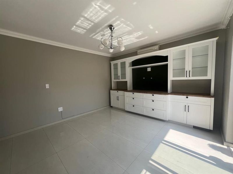 A bright, empty living room featuring large-format floor tiles, neutral grey walls, and extensive white built-in cabinetry with glass display sections.