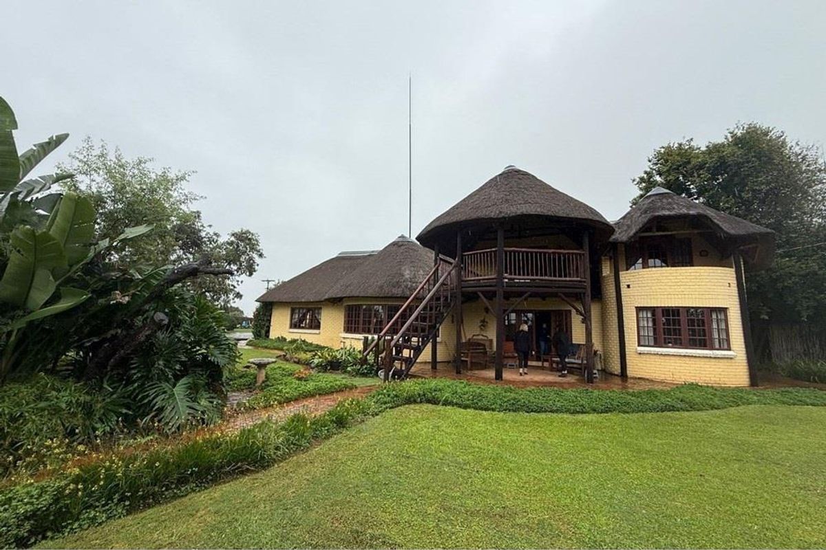 Rear view of a charming thatch-roofed house featuring a timber balcony, external staircase, and a well-maintained garden with a paved patio area.