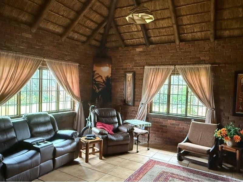 A rustic living room featuring exposed brick walls, a high thatched ceiling with timber beams, and tiled flooring with large windows.