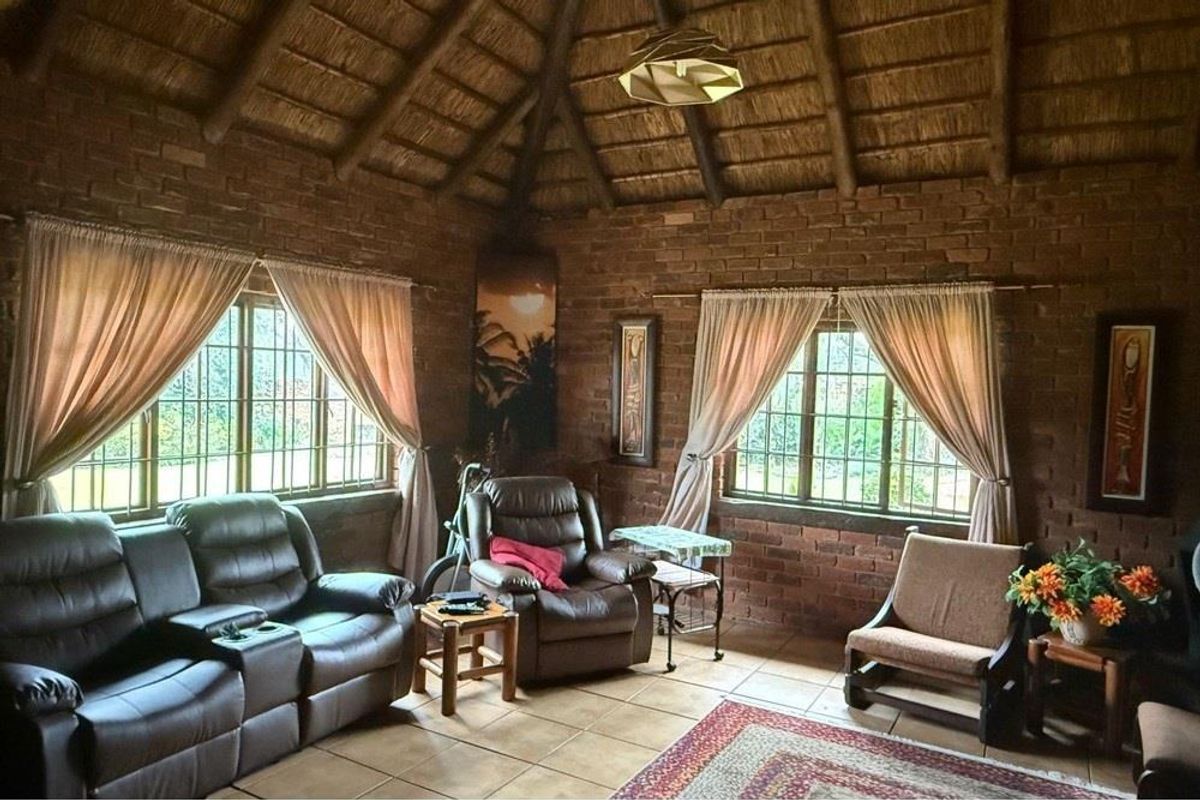 A rustic living room featuring exposed brick walls, a high thatched ceiling with timber beams, and tiled flooring with large windows.