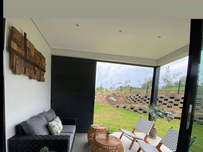 A covered patio area featuring tiled flooring, recessed lighting, and sliding glass doors, overlooking a landscaped garden with a tiered retaining wall.