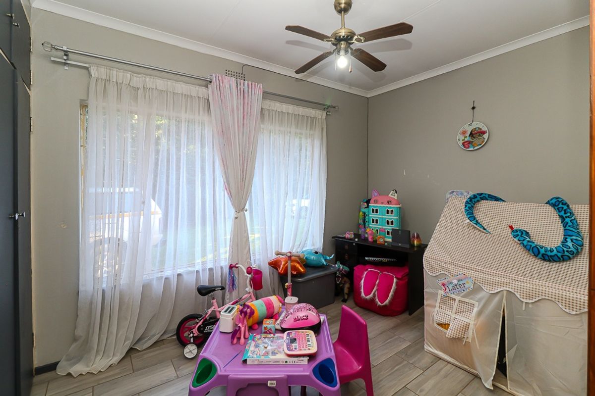 A functional bedroom featuring tiled flooring, a ceiling fan, and large windows with curtain rails, offering a versatile space for various uses.