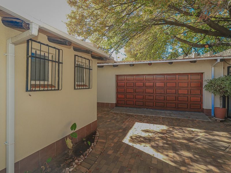 A well-maintained rear exterior area featuring a brick-paved driveway, a wooden garage door, and windows fitted with security bars.
