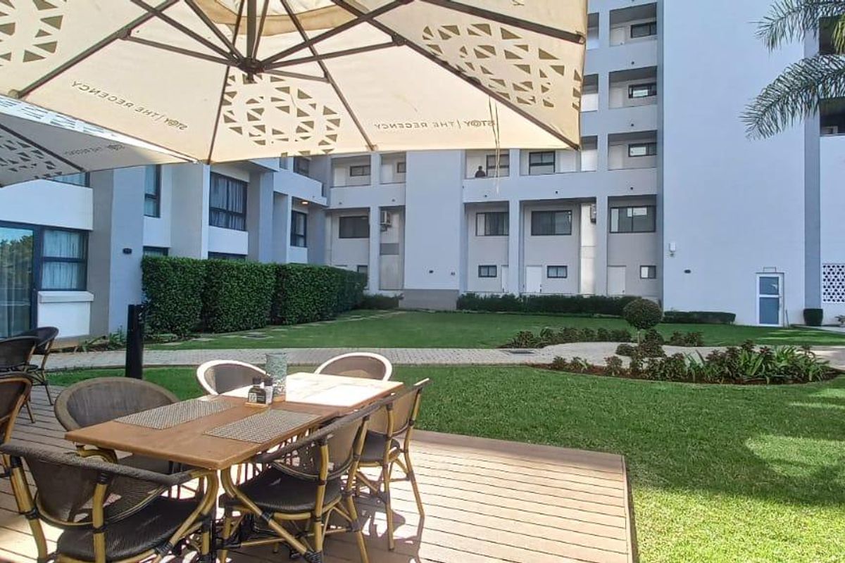 A well-maintained communal garden area featuring timber decking, manicured lawns, and landscaped hedging within an apartment complex courtyard.