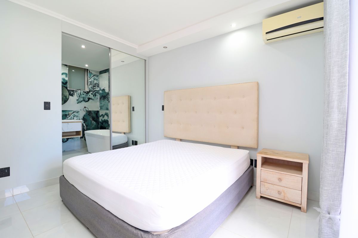A modern bedroom featuring tiled flooring, recessed ceiling lighting, a wall-mounted air conditioning unit, and mirrored sliding wardrobe doors leading to an en-suite bathroom.