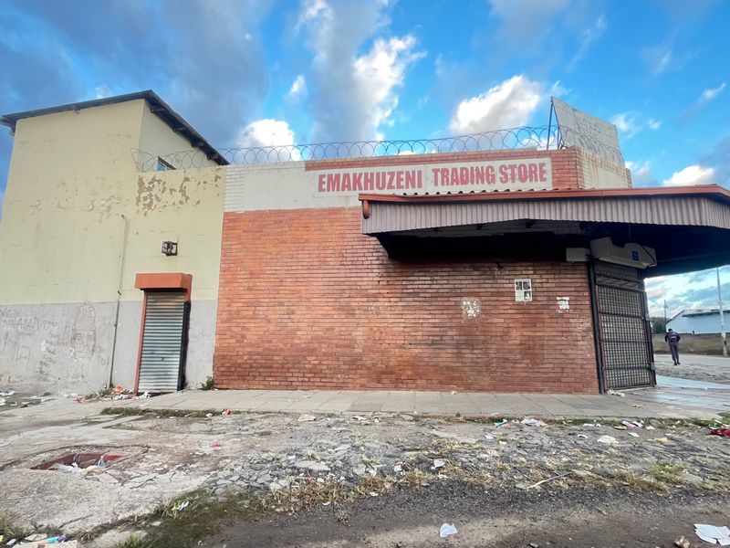 Front elevation of a commercial trading store featuring brick construction, metal roller shutters, and a covered entrance area with perimeter security fencing.