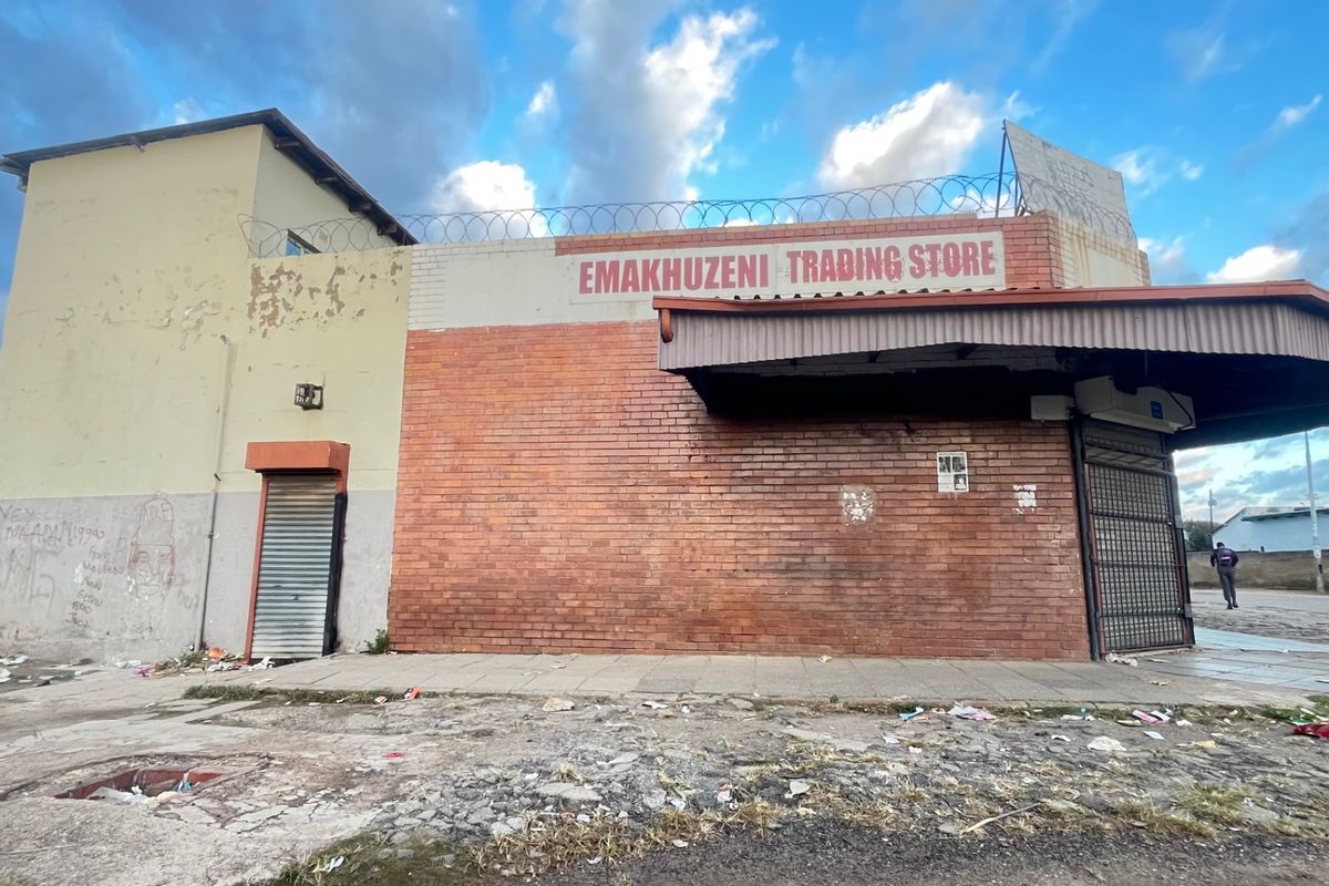 Front elevation of a commercial trading store featuring brick construction, metal roller shutters, and a covered entrance area with perimeter security fencing.