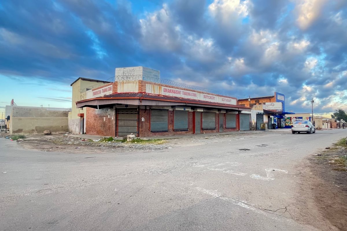 Street-level view of a commercial corner property featuring a brick facade, security shutters, and protective razor wire fencing.