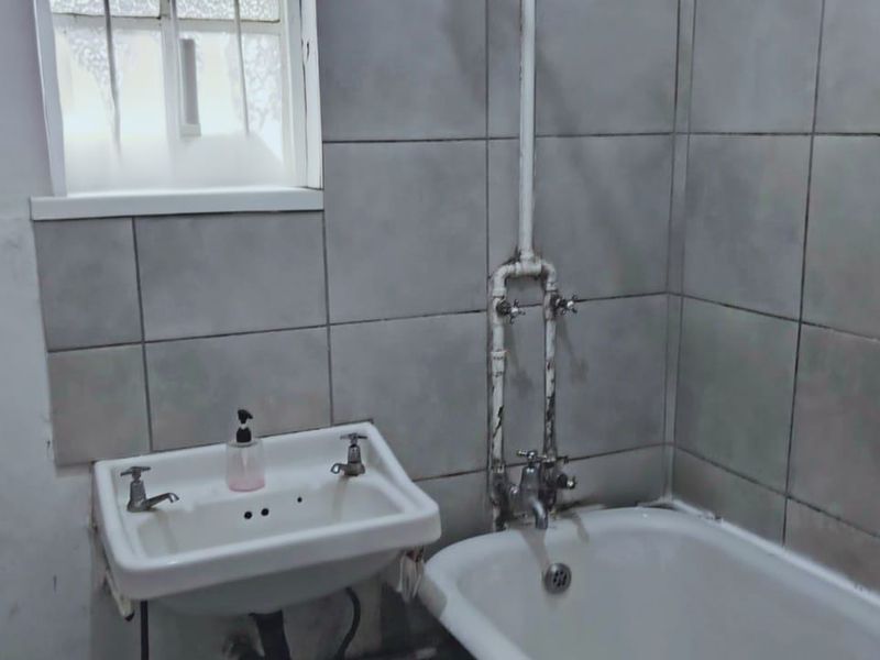 A dated bathroom featuring a wall-mounted basin, bathtub, and exposed plumbing, requiring cosmetic updates and repairs to the wall finishes.