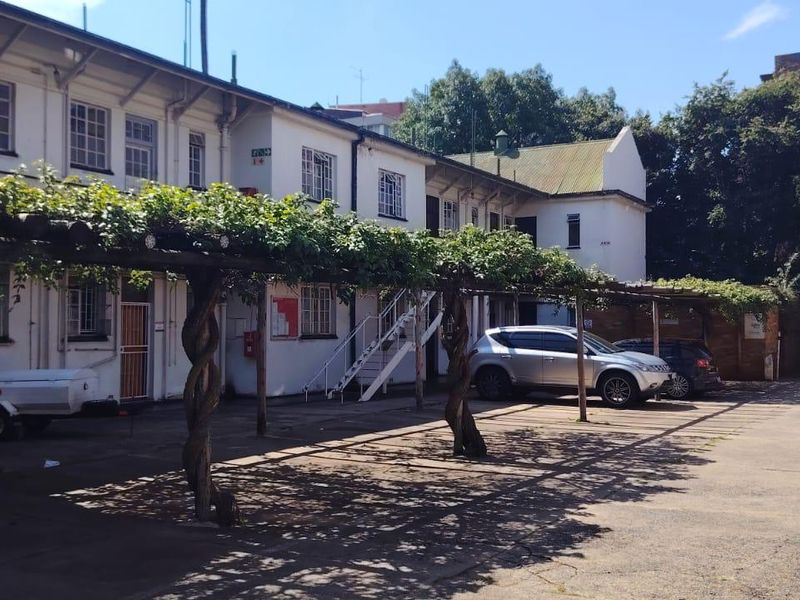 Rear exterior view of the apartment complex featuring a paved parking area with a shaded pergola structure and external staircase access.