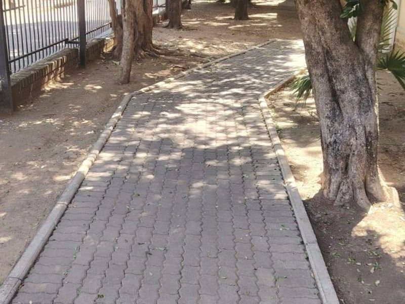 A paved walkway leads through a shaded garden area featuring mature trees and a secure metal perimeter fence.