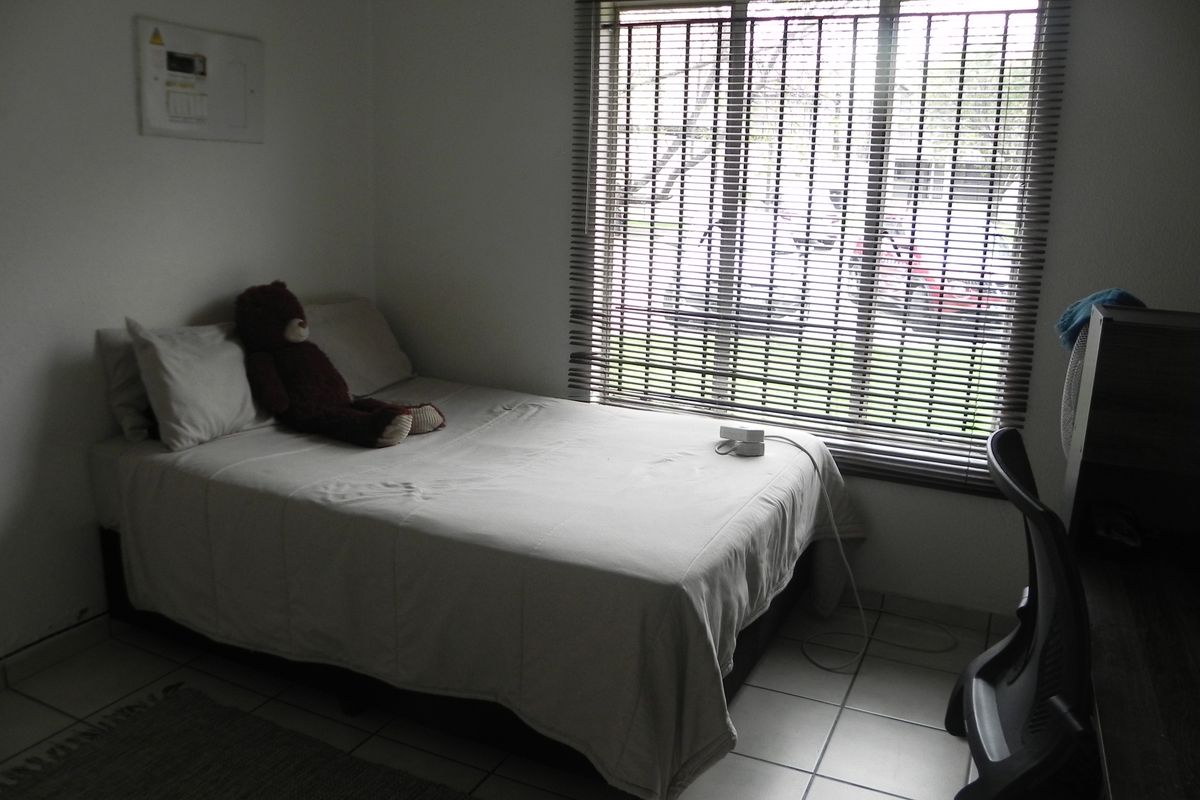 A functional bedroom featuring tiled flooring, a large window with fitted blinds, and a wall-mounted electrical distribution board.