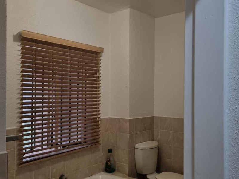 A functional bathroom featuring a built-in bathtub with tiled surround, a standard toilet, and wooden venetian blinds.