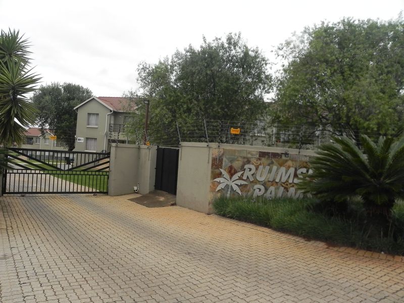 Entrance view of Ruimsig Palms complex featuring a paved driveway, secure gated access, and perimeter walling with electric fencing.