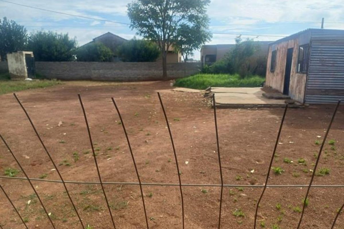Vacant residential land featuring a perimeter wall, wire fencing, and a small corrugated metal structure with an adjacent concrete slab.