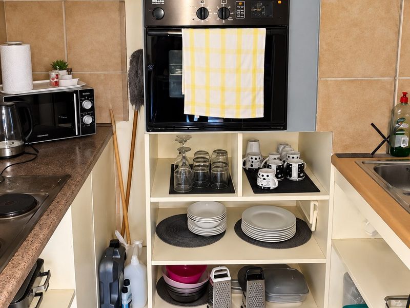 Compact kitchen featuring a built-in Miele oven, ceramic hob, tiled backsplash, and open shelving storage with neutral floor tiling.