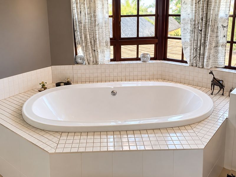 A well-maintained bathroom featuring a large built-in oval bathtub set within a tiled surround, positioned beneath a bright bay window.