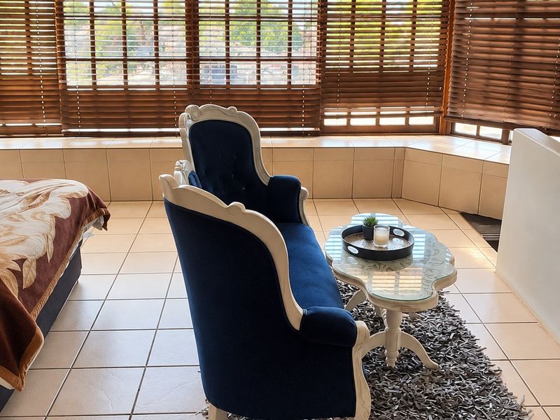 A bright living area featuring large bay windows with venetian blinds and neutral tiled flooring, offering a clean and functional space.