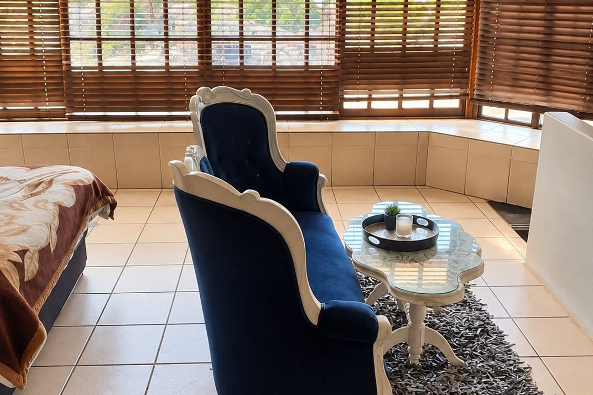 A bright living area featuring large bay windows with venetian blinds and neutral tiled flooring, offering a clean and functional space.