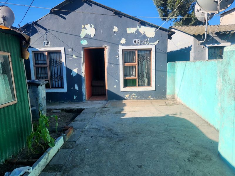 Rear exterior view of a house featuring a concrete paved yard, security gates on windows, and a boundary wall, showing significant exterior paint deterioration.