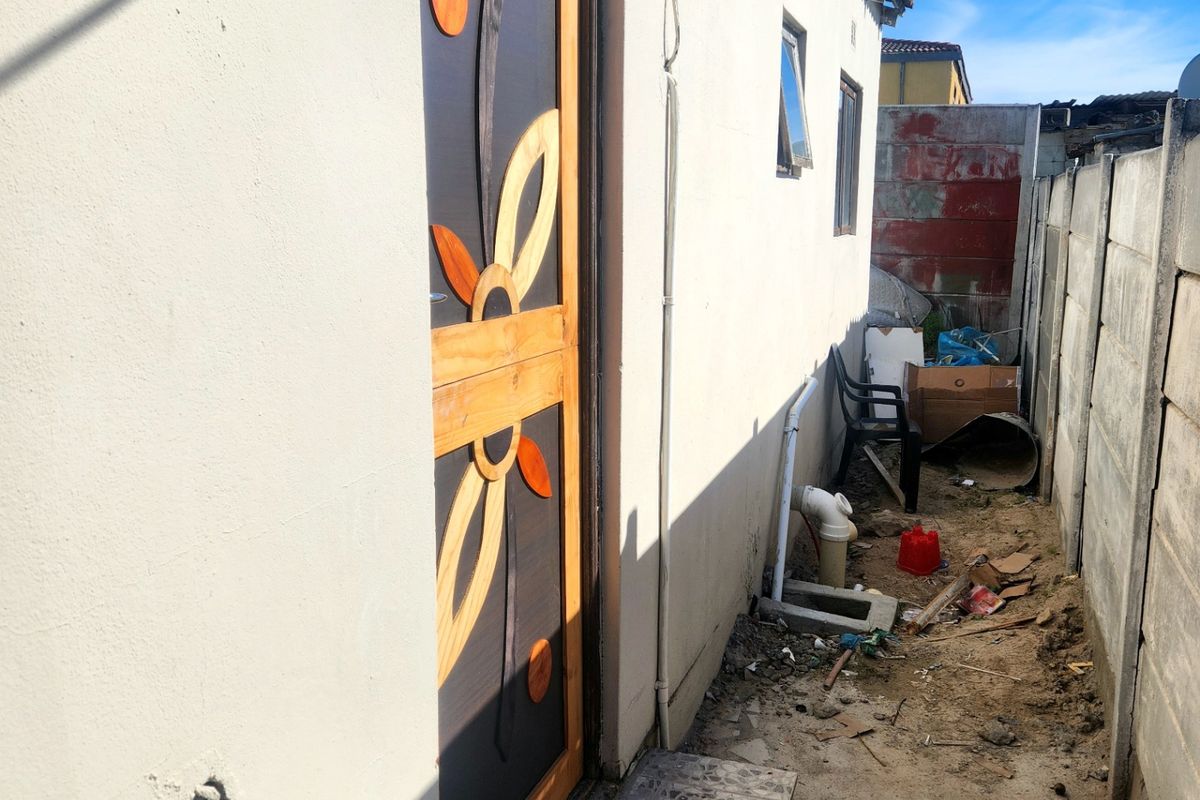 A narrow rear exterior passage featuring a decorative security door, tiled step, and precast concrete boundary wall with unpaved ground.