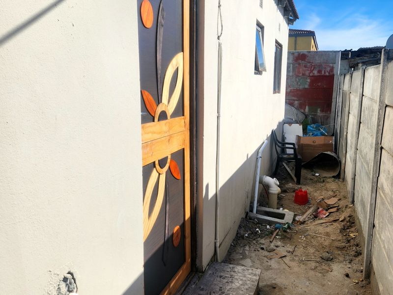A narrow rear exterior passage featuring a decorative security door, tiled step, and precast concrete boundary wall with unpaved ground.
