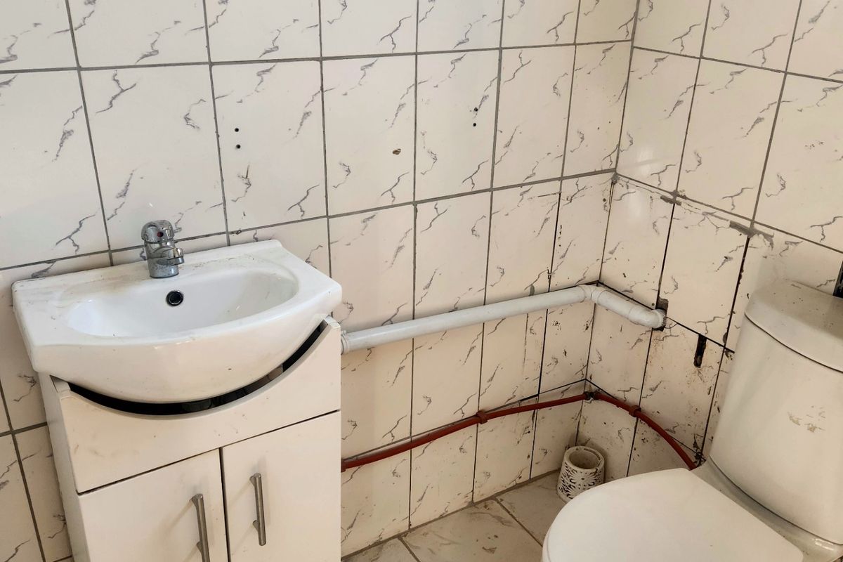 A small bathroom featuring a vanity basin and toilet with visible surface-mounted plumbing and dated wall tiling requiring maintenance or renovation.