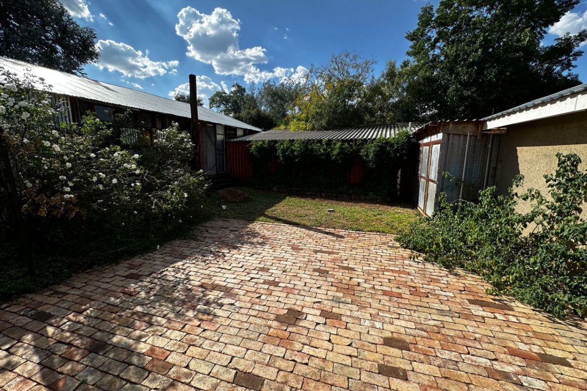 A rear exterior view featuring a brick-paved patio area, corrugated metal roofing, and surrounding garden space with mature trees.