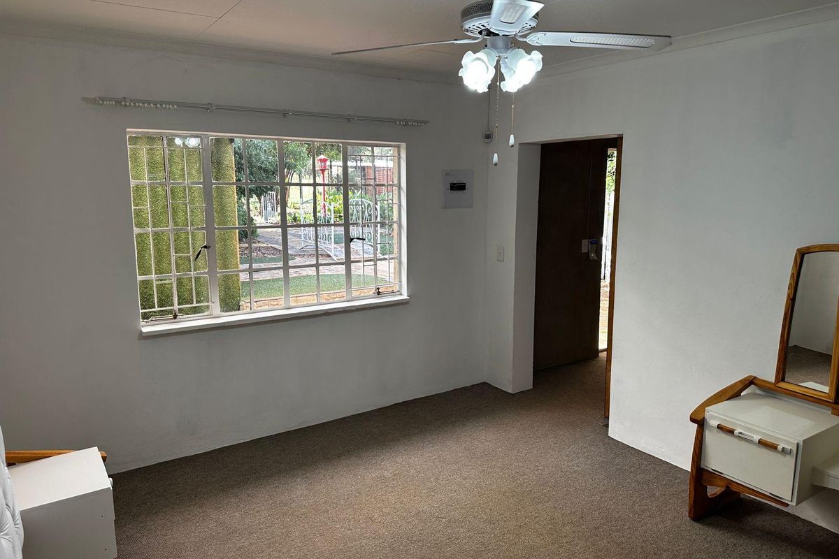 A bedroom featuring carpeted flooring, a ceiling fan, and a window with security burglar bars, offering a functional and neutral living space.