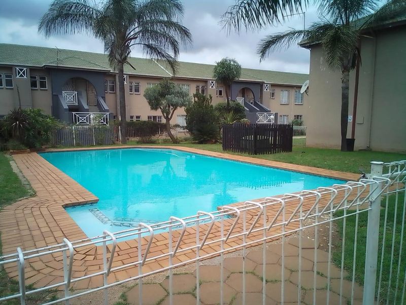 Communal swimming pool area featuring brick coping, metal perimeter fencing, and surrounding landscaped garden within an apartment complex setting.