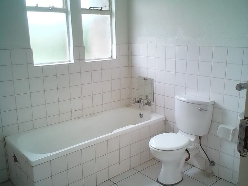 A dated bathroom featuring a standard bathtub, toilet, and white tiled finishes, requiring cosmetic updates and repairs to the bathtub apron.