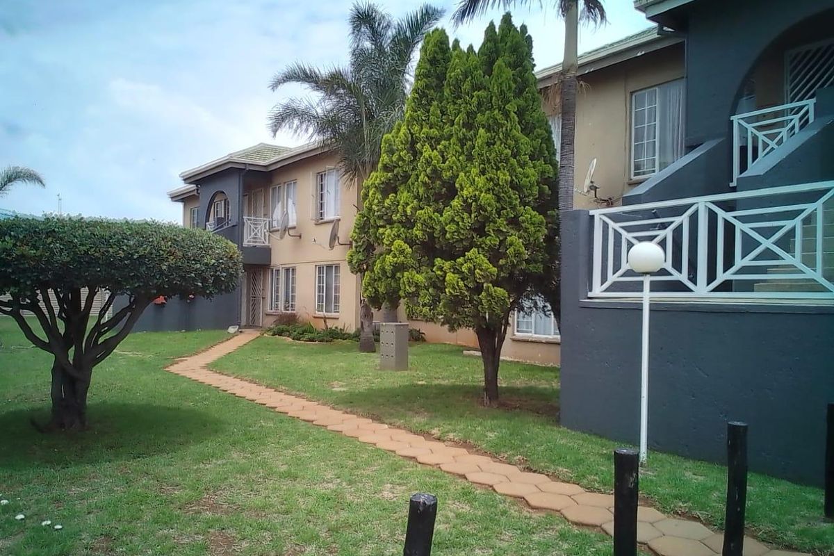 A view of the apartment complex exterior featuring a paved garden walkway, landscaped lawn, and private balcony access.
