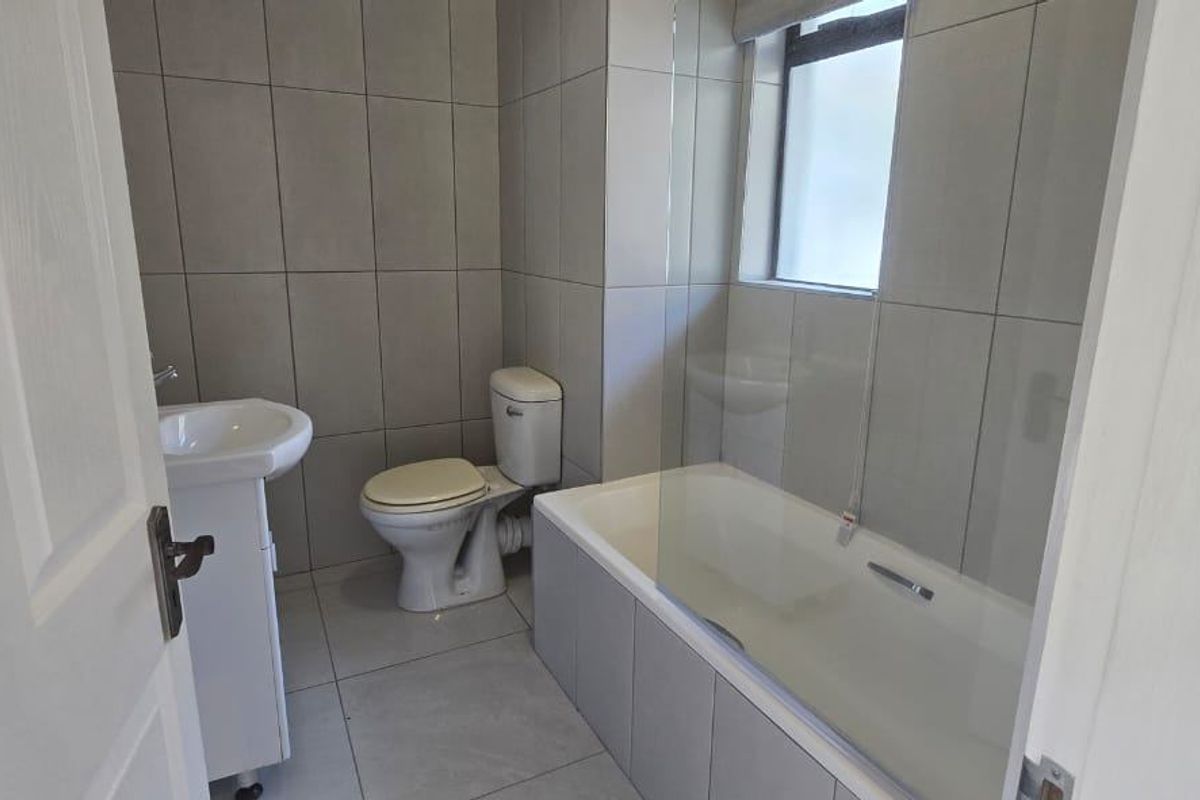 A clean, modern bathroom featuring a pedestal basin, toilet, and a bathtub with a glass shower screen, finished with neutral wall and floor tiles.