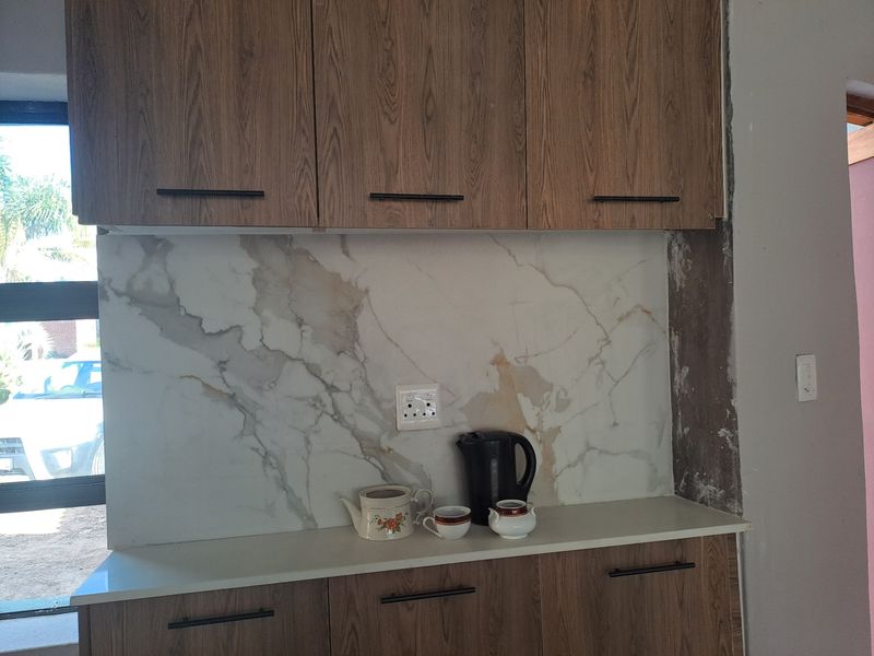 A kitchen area featuring wood-grain cabinetry, a marble-look splashback, and stone countertops, currently showing some unfinished wall edges.