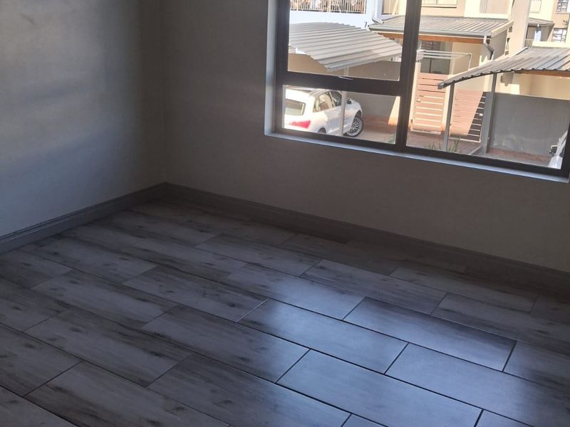 A bright, empty bedroom featuring modern wood-look tiled flooring and a large aluminium-framed window with views of the complex exterior.