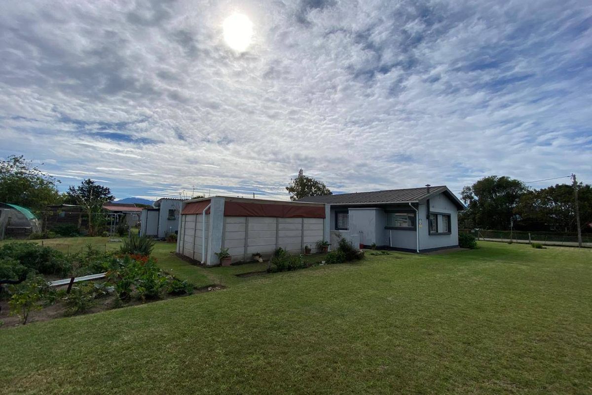 Rear view of the property showing a single-story house with a metal roof, adjacent precast walling, and a large surrounding lawn.