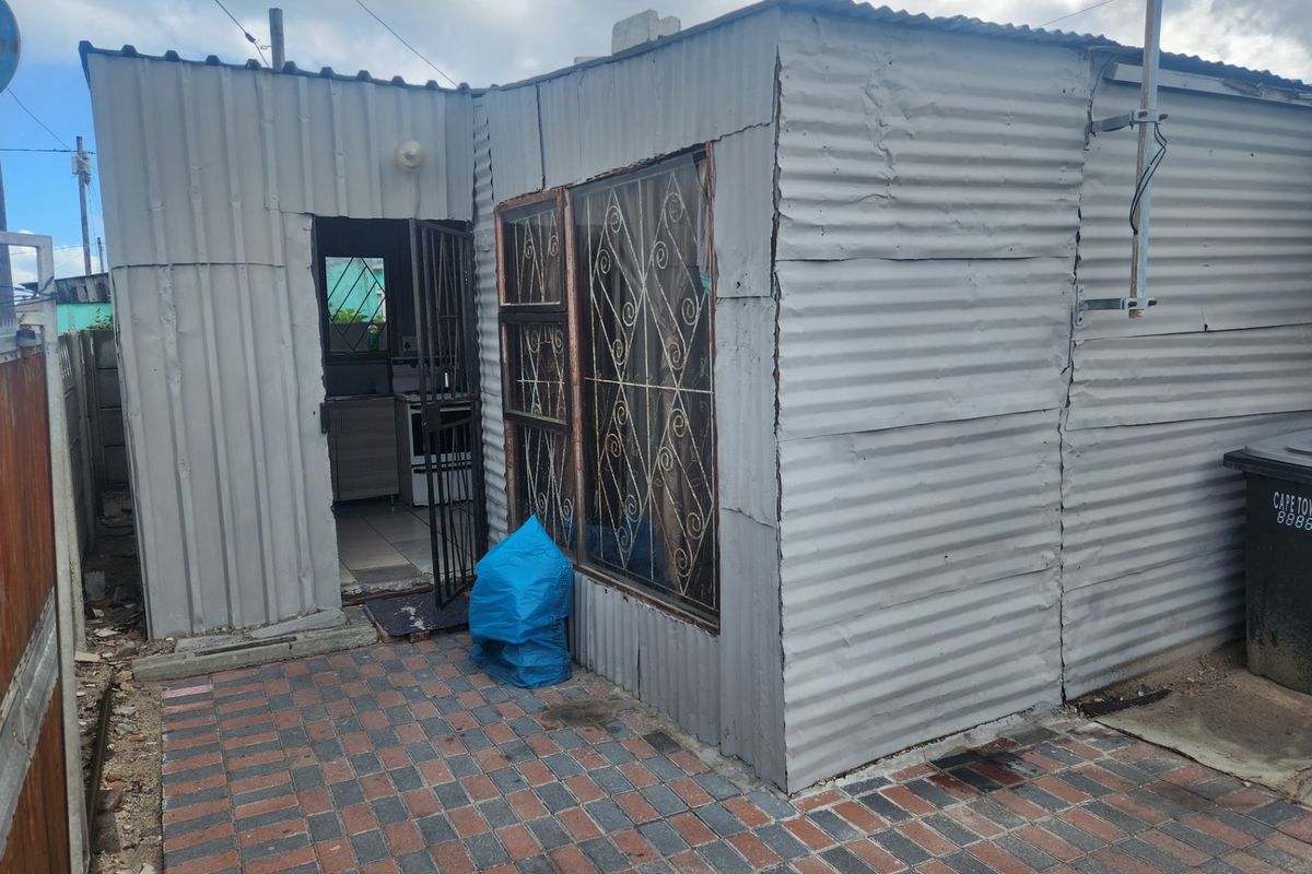 Exterior view of a small residential structure featuring corrugated metal cladding, security bars on windows, and a paved entrance area.