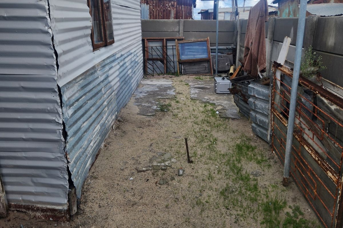 A narrow rear exterior area featuring corrugated metal siding on the structure and a precast concrete boundary wall on a sandy surface.