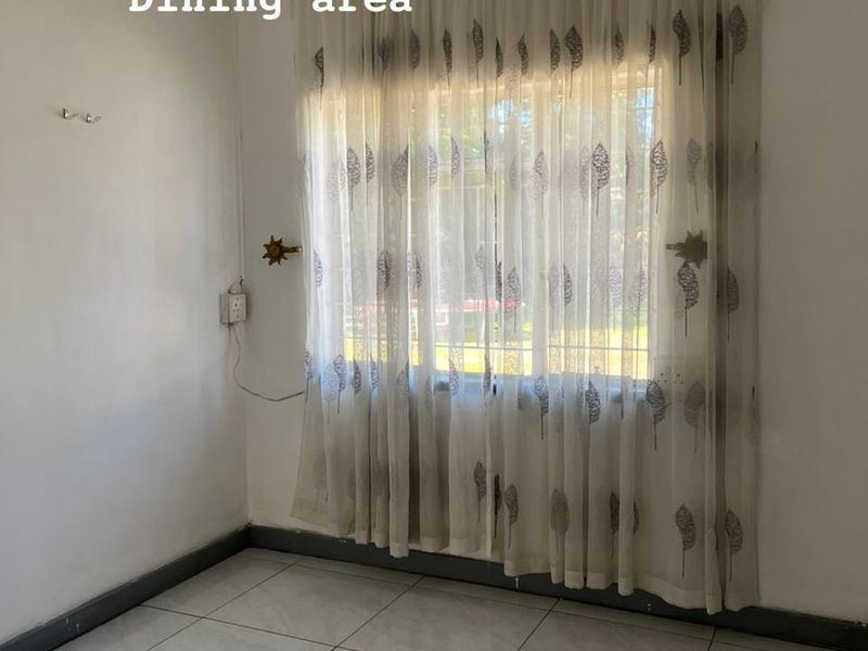A bright, empty dining area featuring tiled flooring, neutral walls, and a window fitted with security bars and decorative curtains.